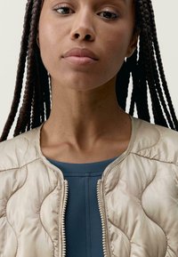 Světle béžová prošívaná bomber bunda s metalickým zipem, nošená přes tmavě modrý přiléhavý top. Materiál obsahuje jemný vlnkový vzor.