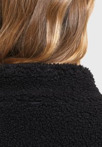 Cheveux châtain clair mi-longs reposant sur le col d'une veste polaire noire texturée sur un fond uni.