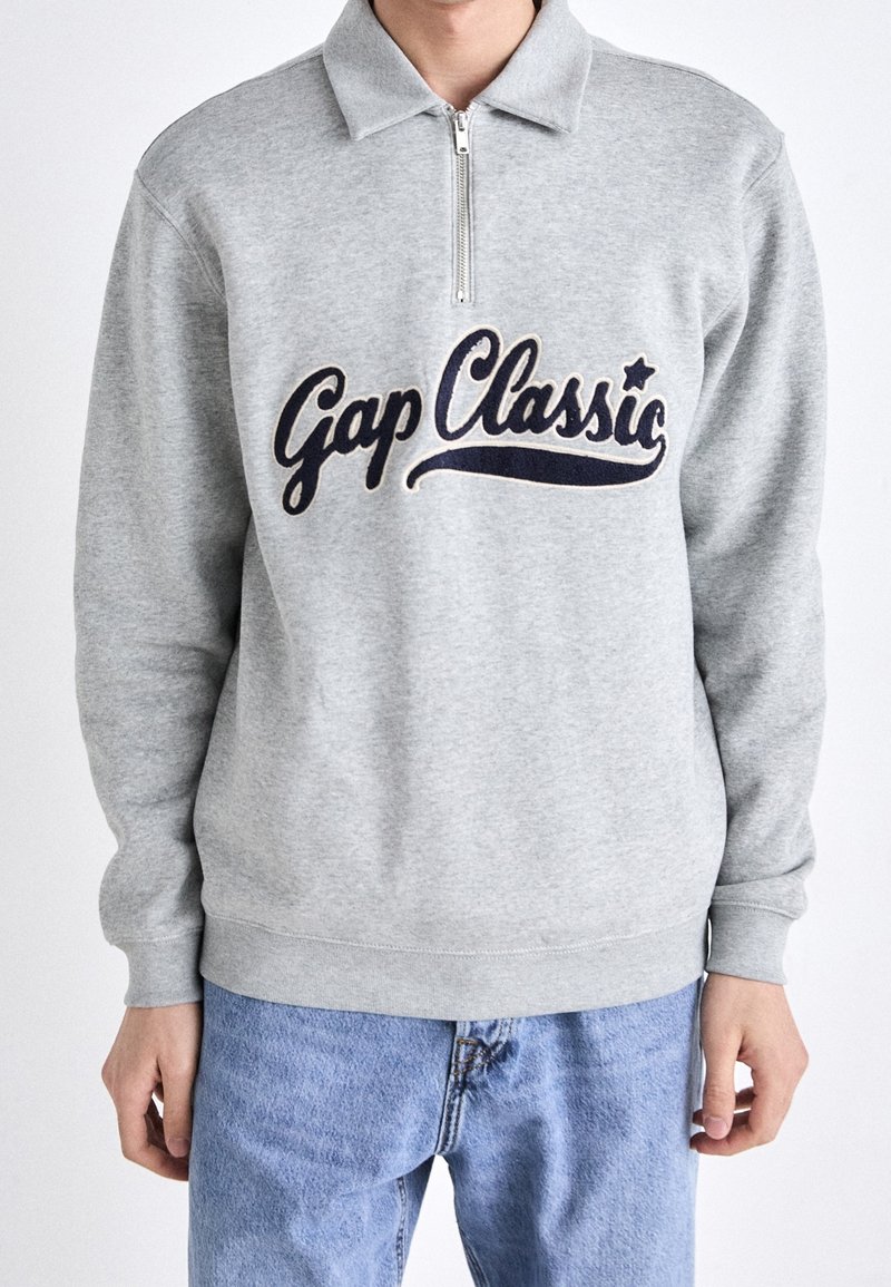 Sweat-shirt gris clair à col zippé quart avec le texte « Gap Classic » en bleu marine, porté avec un jean bleu clair, montrant le torse et les bras.