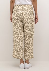 Personne portant un pantalon court à rayures zèbre beige et blanc et des baskets blanches, se tenant devant un fond clair et uni.