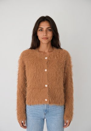 Donna con capelli lunghi e scuri che indossa un cardigan beige peloso abbottonato e jeans azzurri, in piedi davanti a uno sfondo chiaro e uniforme.