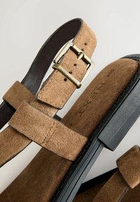 Sandales en daim marron avec bride à boucle réglable, semelle plate noire et texture lisse. Couture remarquable le long des bords.