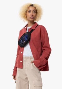 Červená kostkovaná nadměrná košile, šedý crew neck top pod tím, béžové cargo kalhoty a černá taška přes rameno s modrými akcenty.