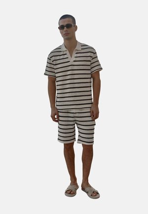 Mand iført sort-hvid stribet kortærmet striktrøje og matchende shorts med beige slide-sandaler og mørke solbriller.