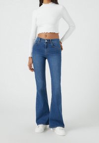 Mujer vistiendo una blusa blanca de manga larga corta, jeans acampanados azul de cintura alta, y zapatillas blancas, de pie frente a un fondo sencillo.