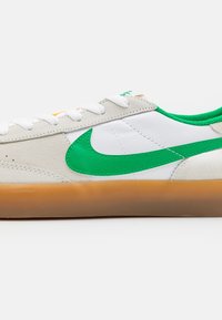 Nízké bílé tenisky s béžovými semišovými akcenty, zeleným Nike swoosh a vnitřní podšívkou, bílými tkaničkami a gumovou podrážkou.