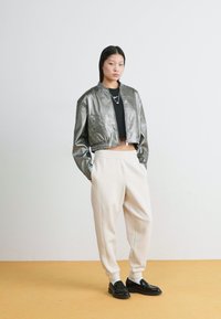 Metalna srebrna krajša bombaška jakna z dolgimi rokavi, v kombinaciji s kremnimi trenerkami in črnimi loaferji, na nevtralni podlagi.