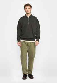 Donkergroene pullover met een ritskraag, voorzien van een subtiele textuur. Gecombineerd met licht olijfgroene broeken. Schoenen zijn donker en glanzend.