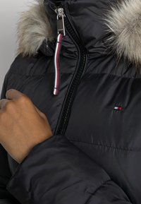 Chaqueta negra tipo plumas con capucha forrada de piel, cremallera plateada y cordón de acento en rojo, blanco y azul. Presenta un parche de logo en el pecho.