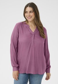 Kobieta uśmiechająca się, ubrana w długosleeve'owy bluzkę w kolorze mauve z guzikami i niebieskie dżinsy, stojąca na jednolitym białym tle.