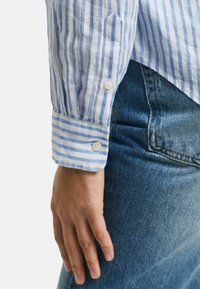 Camicia a righe blu e bianche con bottoni e polsini, abbinata a jeans in denim blu. Il tessuto appare leggero e strutturato.