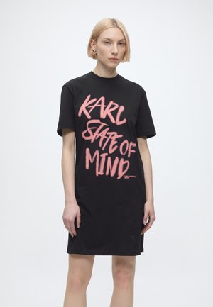 Femme blonde portant une robe noire à manches courtes avec un texte rose "KARL STATE OF MIND" sur le devant, sur un fond blanc uni.