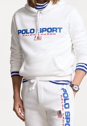 Persoon draagt een witte Polo Sport Ralph Lauren hoodie en bijpassende joggingbroek met blauw en rood logo en details van de Amerikaanse vlag.