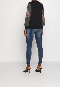 Kvinna som står med ryggen vänd mot kameran, bärande en genomskinlig svart blus med blommiga ärmar, blå slim fit-jeans och svarta högklackade mules.