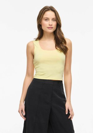 Donna con capelli castani lunghi che indossa un top giallo senza maniche e pantaloni neri a vita alta, in piedi davanti a uno sfondo bianco.