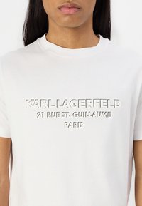 Balta medvilninė marškinėliai su reljefiniu užrašu "KARL LAGERFELD 21 RUE ST-GUILLAUME PARIS" priekyje. Šikia kaklo dizainas, trumpi rankovės.