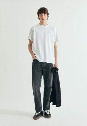 Junger Mann steht, trägt ein weißes T-Shirt und schwarze Jeans, hält eine schwarze Jacke in einer Hand vor einem einfarbigen hellen Hintergrund.