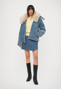 Mujer vestida con una chaqueta de pelo azul, suéter amarillo abotonado, falda azul y botas negras hasta la rodilla, de pie contra un fondo blanco.