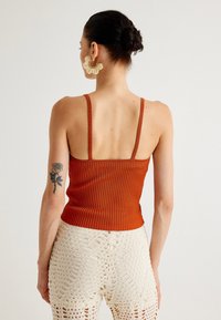 Top arancione a righe con spalline sottili, abbinato a pantaloni bianchi all'uncinetto con motivi traforati. Orecchini a dichiarazione floreali dorati.