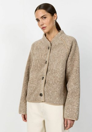 Sieviete ar tumšiem matiem zemas zirgastes frizūrā, valkājot bēša krāsas teksturētu, ar pogām aizvērtu žaketi un gaismas krāsas bikses, stāvot pret vienkāršu fona sienu.