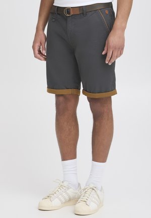 Shorts gris avec des revers marron, des poches avant, une ceinture marron et une coupe décontractée. Portés avec des baskets blanches et des chaussettes.