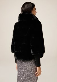 Mujer con cabello oscuro que lleva una chaqueta de pelaje sintético negro y una falda de lentejuelas plateadas, de perfil frente a un fondo sencillo.