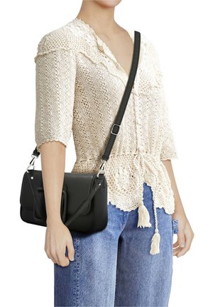 Femme portant un haut en crochet crème et un jean bleu, avec un sac bandoulière en cuir noir doté d'une sangle réglable et d'une poche avant.
