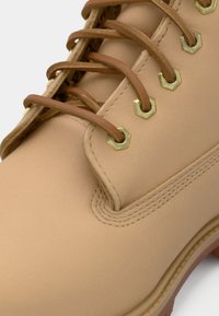 Gros plan sur une botte en cuir beige avec des lacets marron passant par des œillets hexagonaux dorés et des détails de couture visibles.