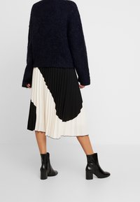 Osoba ubrana w czarny puszysty sweter, czarną i białą plisowaną spódnicę midi oraz czarne botki na obcasie, stojąca na szarym podłożu.