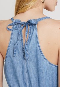 Femme blonde portant une robe en denim sans manches avec un nœud noué à l'encolure arrière, dévoilant le haut de son dos et son épaule.