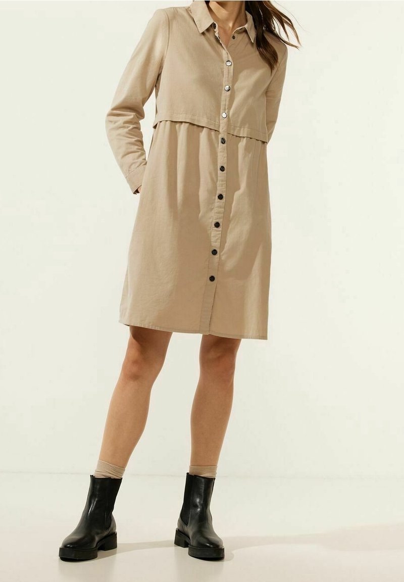 Robe beige boutonnée avec manches longues, col chemise et corsage ajusté s'évasant en une jupe ample. Boutons noirs et bottines noires.