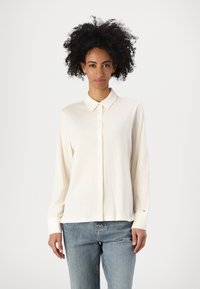 Kremfarget skjorte med knapper laget av mykt stoff, har klassisk krage, lange ermer og en avslappet passform, kombinert med blå jeans.