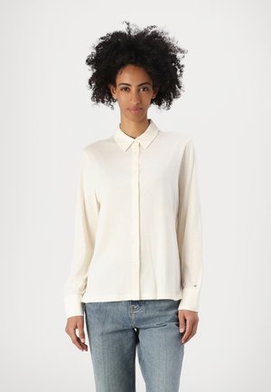 Kremfarget skjorte med knapper laget av mykt stoff, har klassisk krage, lange ermer og en avslappet passform, kombinert med blå jeans.
