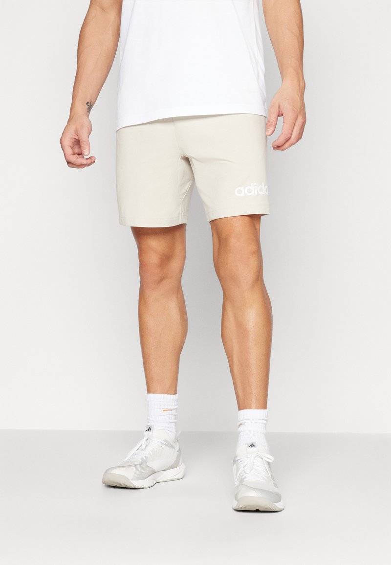 Shorts beiges en mélange de coton avec une taille élastique et un logo Adidas blanc. Portés avec des chaussures de sport blanches et des chaussettes côtelées.