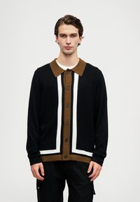 Fiatal férfi fekete, barna és fehér gombos kardigánt visel fehér ing felett, egy sima világosszürke háttér előtt áll.