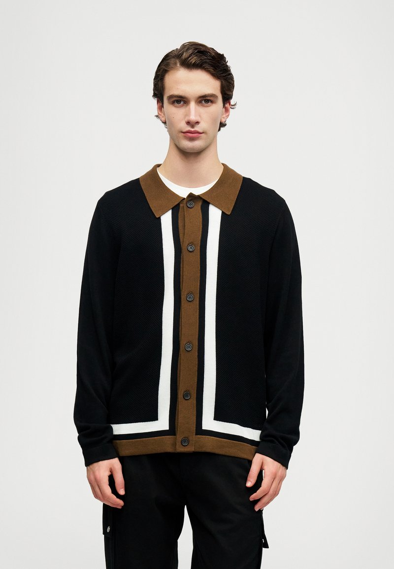 Fiatal férfi fekete, barna és fehér gombos kardigánt visel fehér ing felett, egy sima világosszürke háttér előtt áll.