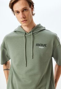 Green short-sleeve hoodie made of soft fabric, featuring a front pocket, drawstring hood, and "MOVE Master" logo on the chest.