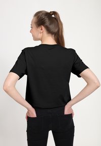 Černé tričko s krátkým rukávem a kulatým výstřihem, z hladkého materiálu a s volným střihem, zobrazené zezadu. Nošeno s černými džínami.