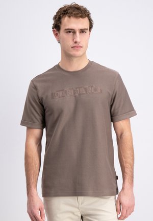 Młody mężczyzna ubrany w gładki brązowy T-shirt z krótkim rękawem, z subtelnym wytłoczonym napisem na piersi, w połączeniu z jasnobeżowymi spodniami, stojący na białym tle.