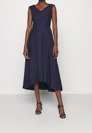 Kobieta w granatowej, bezrękawnikowej sukience z dekoltem w serek i asymetrycznym dołem, czarne sandały na wysokim obcasie z paskiem wokół kostki.