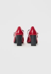 Un paio di scarpe rosse con tacco alto e cinturini alla caviglia, mostrate da dietro su uno sfondo bianco semplice.