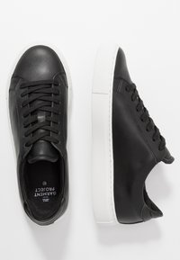 Baskets basses en cuir noir avec semelles épaisses en caoutchouc blanc, lacets noirs et semelles intérieures de marque, présentées en vues de dessus et de côté.