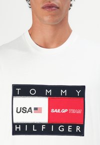Torse masculin portant une chemise blanche avec un grand patch Tommy Hilfiger présentant le texte USA, un petit drapeau et le logo de l'équipe SailGP.