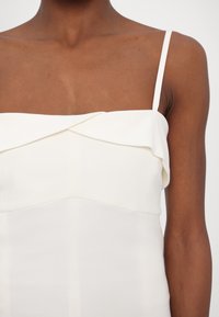 Personne à la peau foncée portant une robe blanche sans manches avec de fines bretelles et un détail de col replié.