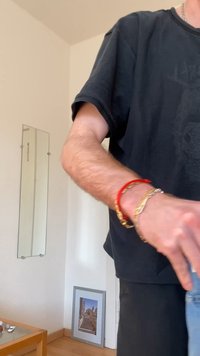 Άτομο που φοράει μαύρο πουκάμισο και βραχιόλια, τεντώνει το χέρι του σε εσωτερικό χώρο κοντά σε έναν τοίχο με έναν μικρό καθρέφτη και μια κορνιζαρισμένη φωτογραφία στο πάτωμα.