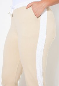 Béžové tepláky s bílou texturovanou bočním panelem a stahovací šňůrkou v pase. Mezi vlastnosti patří kapsy a hladká, jemná textilie.