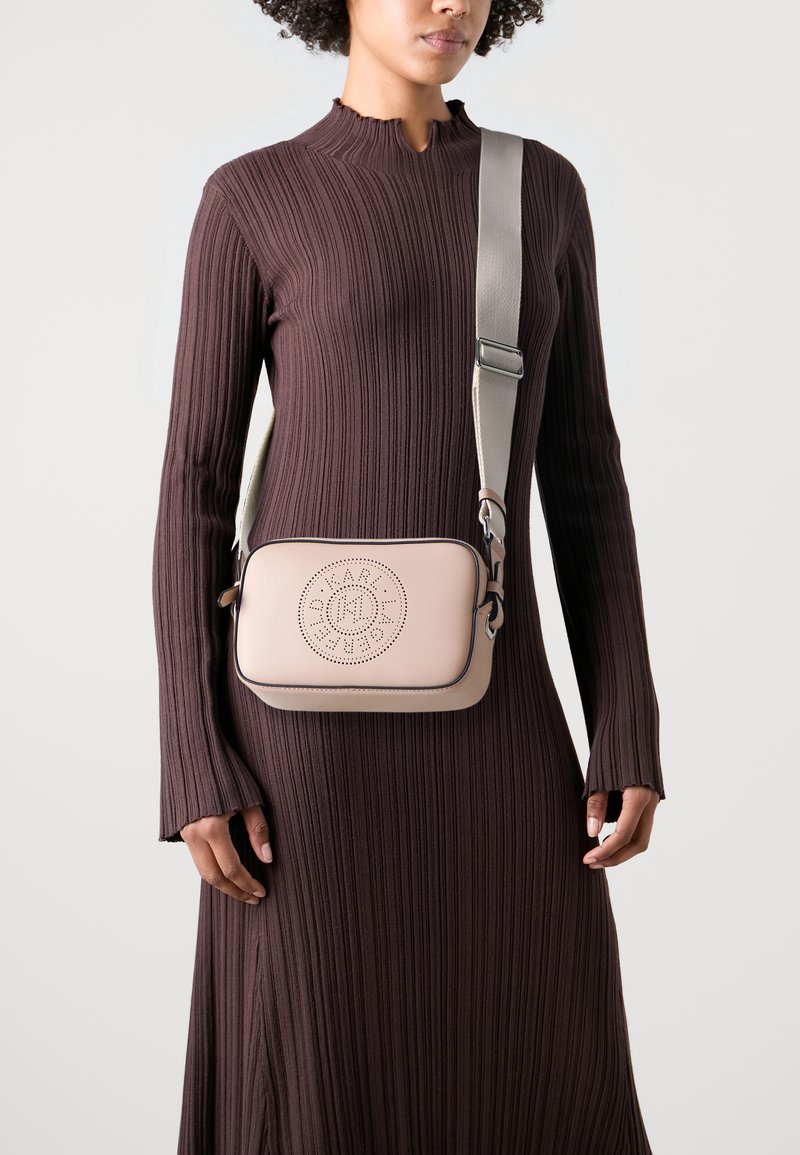 Bežová crossbody taška s perforovaným kruhovým dizajnom, zdobená šedým lemovaním a kovovými doplnkami. Nosí sa cez hnedé šaty s vysokým golierom a s rebrovaným vzorom.
