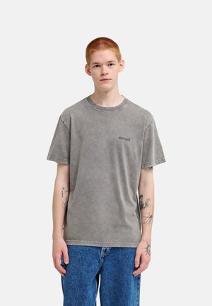 Jeune personne aux cheveux rouges courts portant un t-shirt gris délavé "element" et un jean bleu, debout devant un fond clair uni.