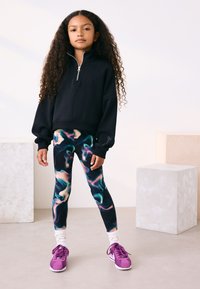 Fekete, cipzáros pulóver túlméretezett ujjal, színes, örvényszerű mintájú, testhezálló leggingsszel párosítva. A szettet lila sportcipő egészíti ki.
