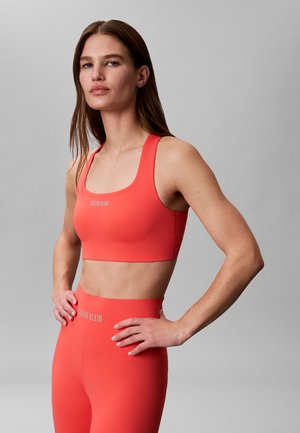 Kobieta o długich brązowych włosach, ubrana w dopasowany koralowy top sportowy i wysokie legginsy, stoi z rękami na biodrach na szarym tle.